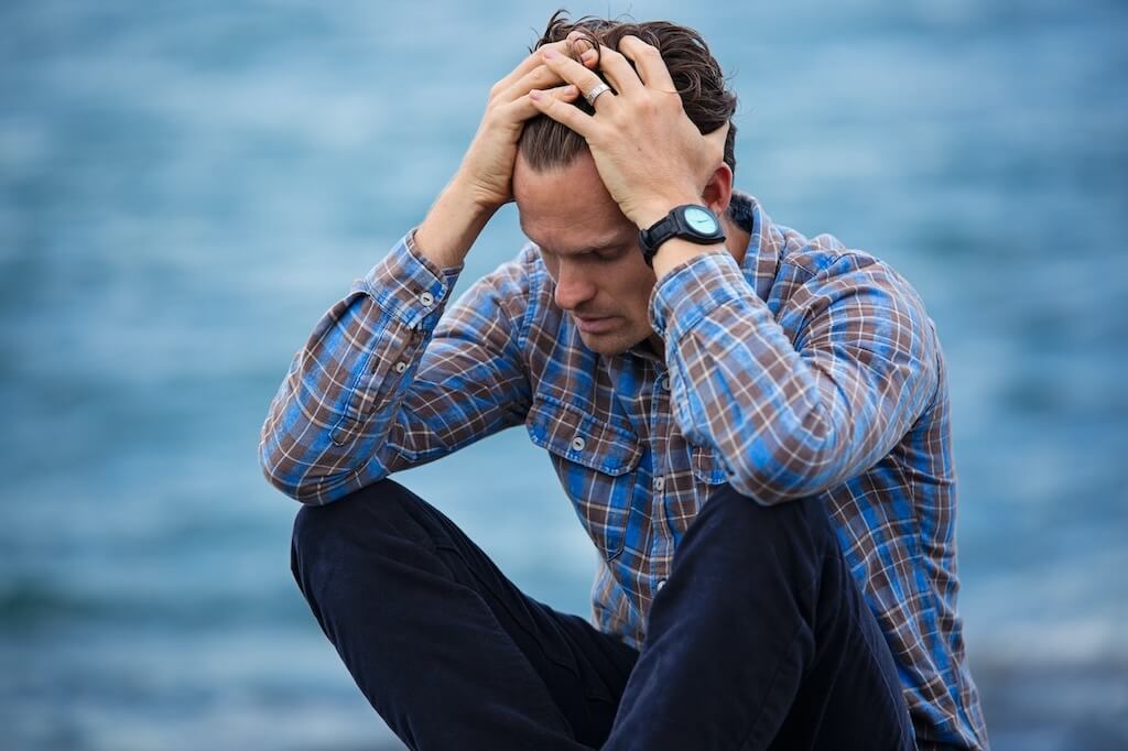 man in brown and blue plaid shirt sitting and holding his head