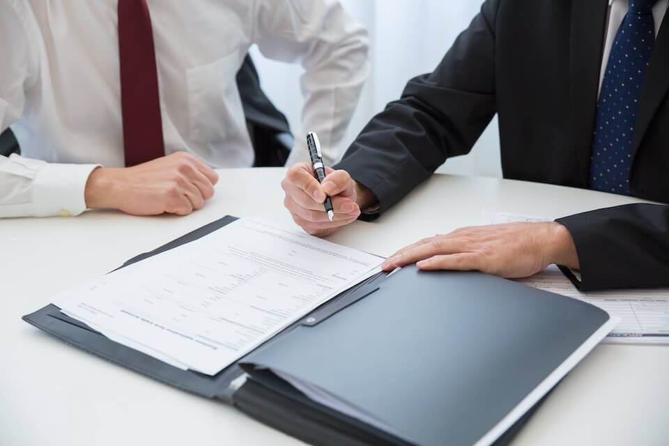 person in black suit preparing to sign a document next to person in red tie