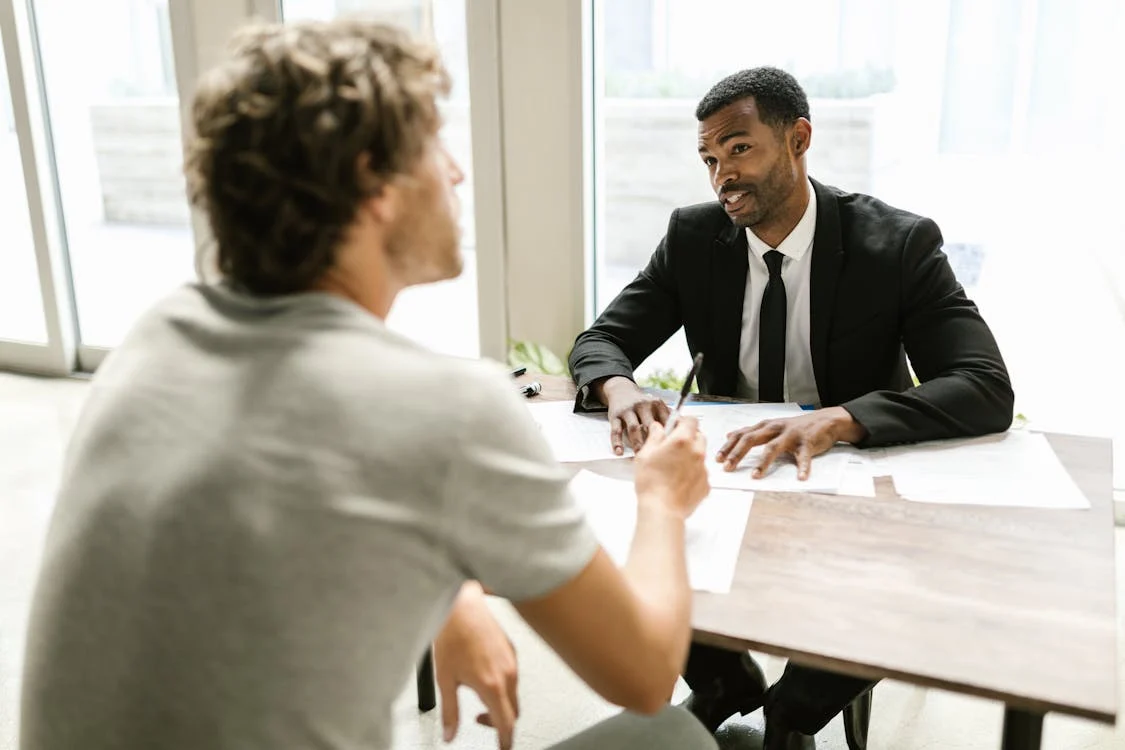 man in t-shirt discussing documents with man in suit at a desk