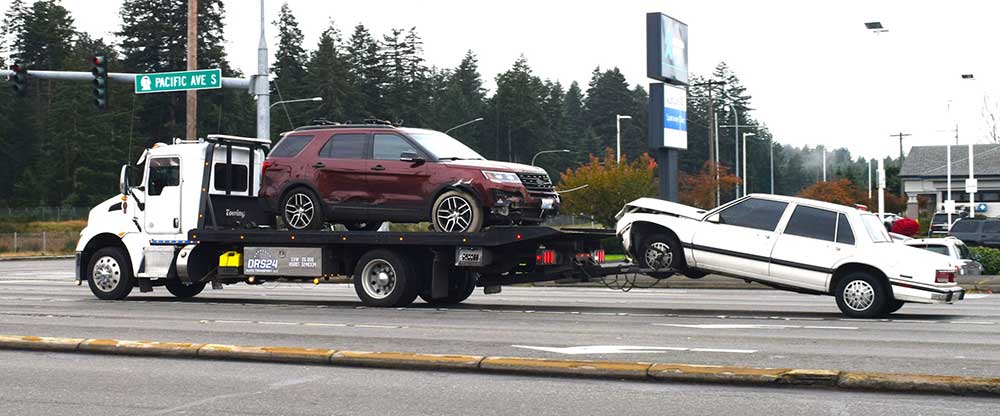 A tow truck driving with two damaged vehicles attached to it
